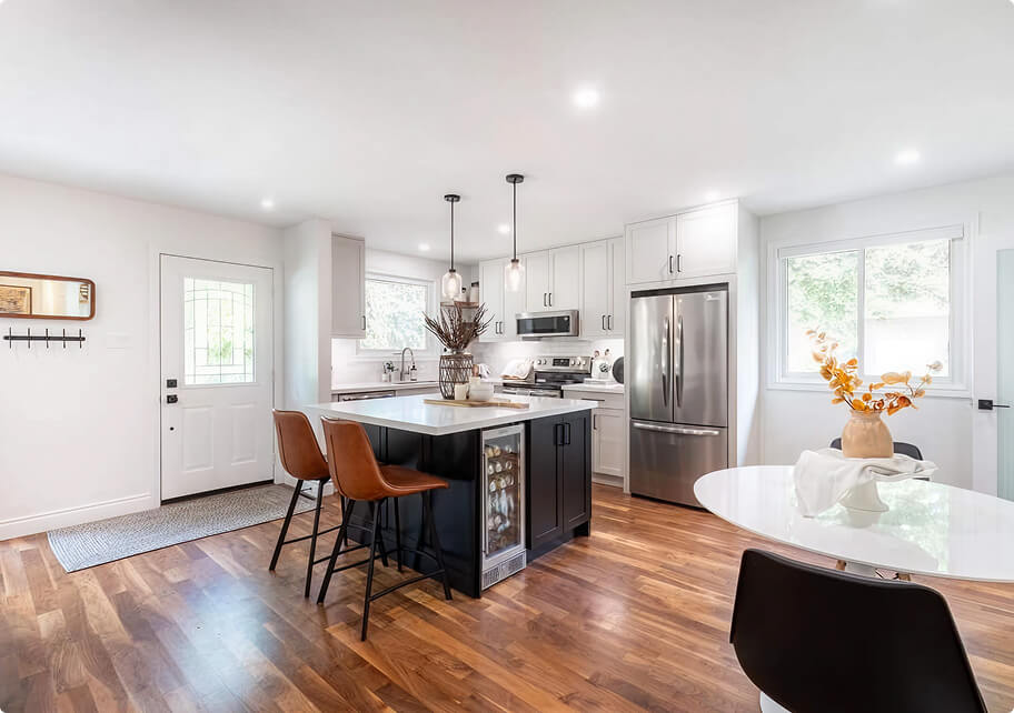 Open-concept kitchen renovation with white upper cabinets, dark contrasting island with wine cooler, leather barstools, hardwood floors, and stainless steel appliances by Odyssey Custom Contracting