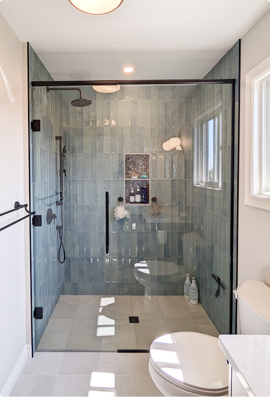 Custom bathroom shower renovation featuring blue-grey subway tile walls, rain showerhead, matte black hardware, mosaic tile niche, and glass door by Odyssey Custom Contracting