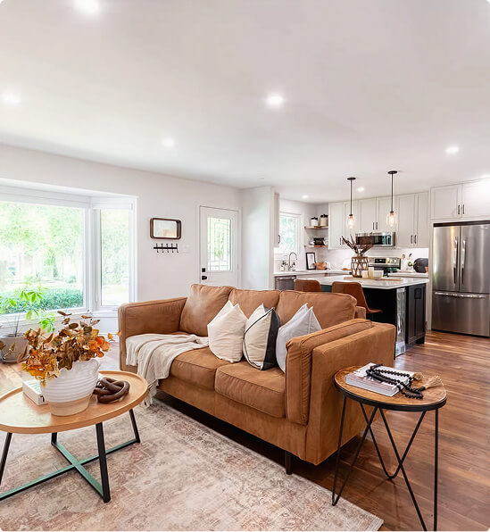 Open-concept home renovation featuring a modern living room with warm caramel sectional sofa, hardwood floors, and a kitchen with dark island and stainless steel appliances by Odyssey Custom Contracting