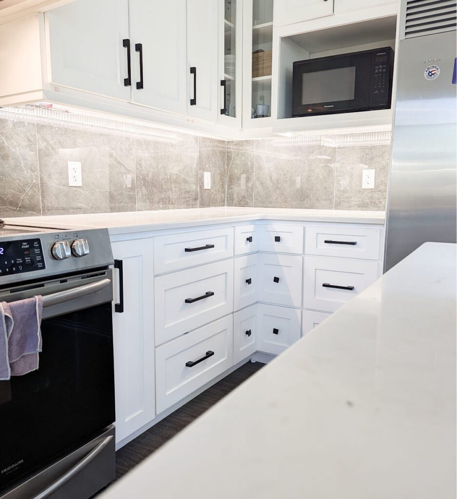 Thoughtfully designed kitchen renovation with white shaker cabinets, black hardware, grey marble tile backsplash, and stainless steel refrigerator by Odyssey Custom Contracting